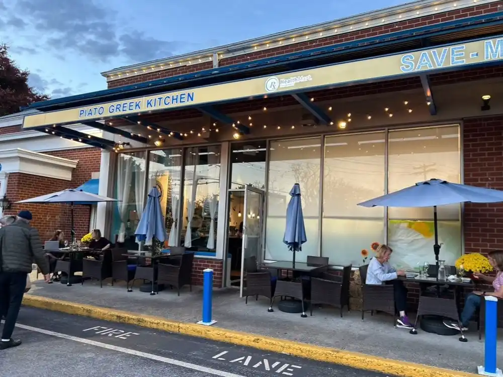 The patio outside Piato Greek Kitchen in the evening, with blue umbrellas, string lights and the blue sign over the windows