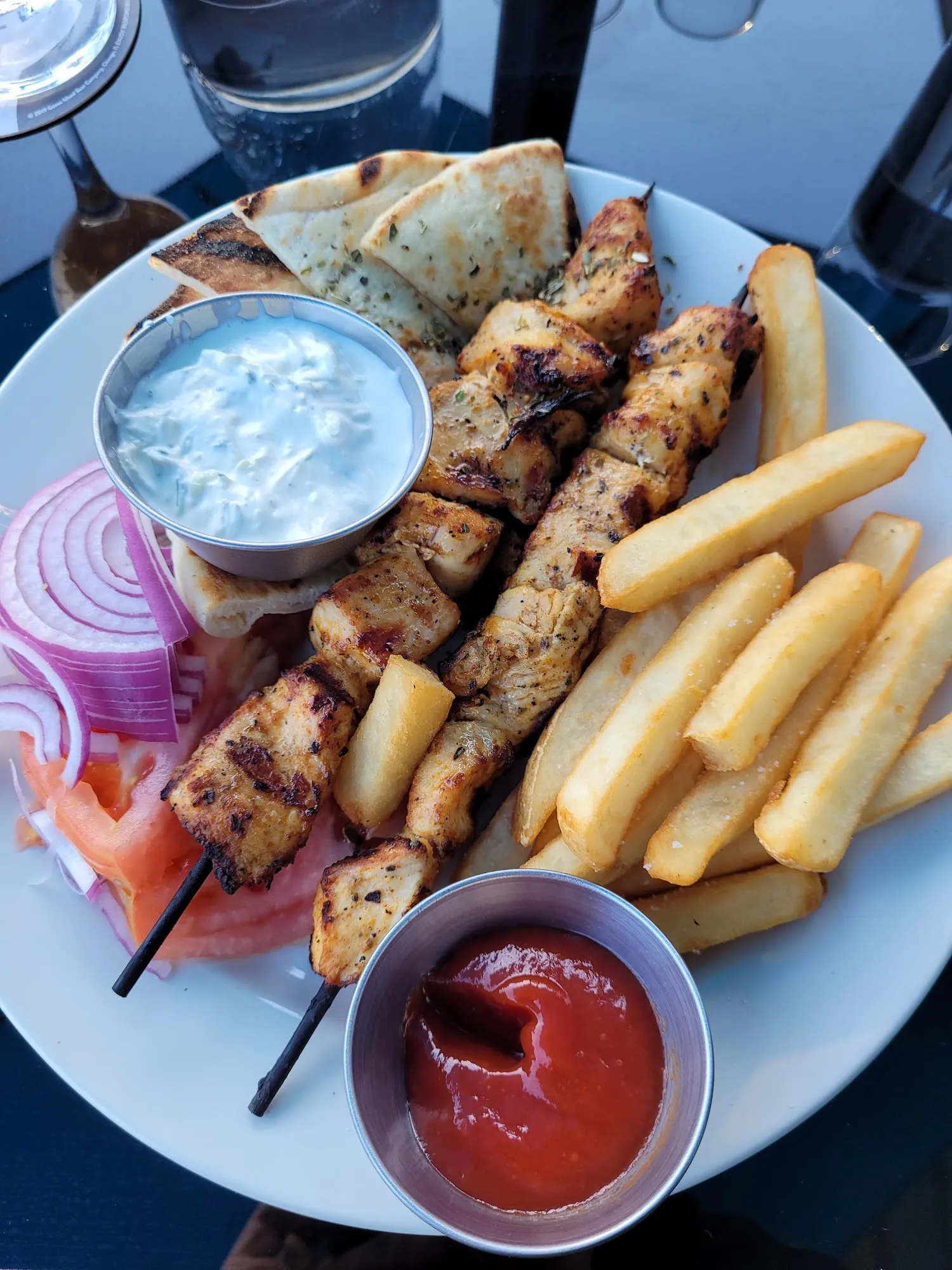 A chicken souvlaki platter with skewers, fresh cut fries, pita, red onion and tzatziki